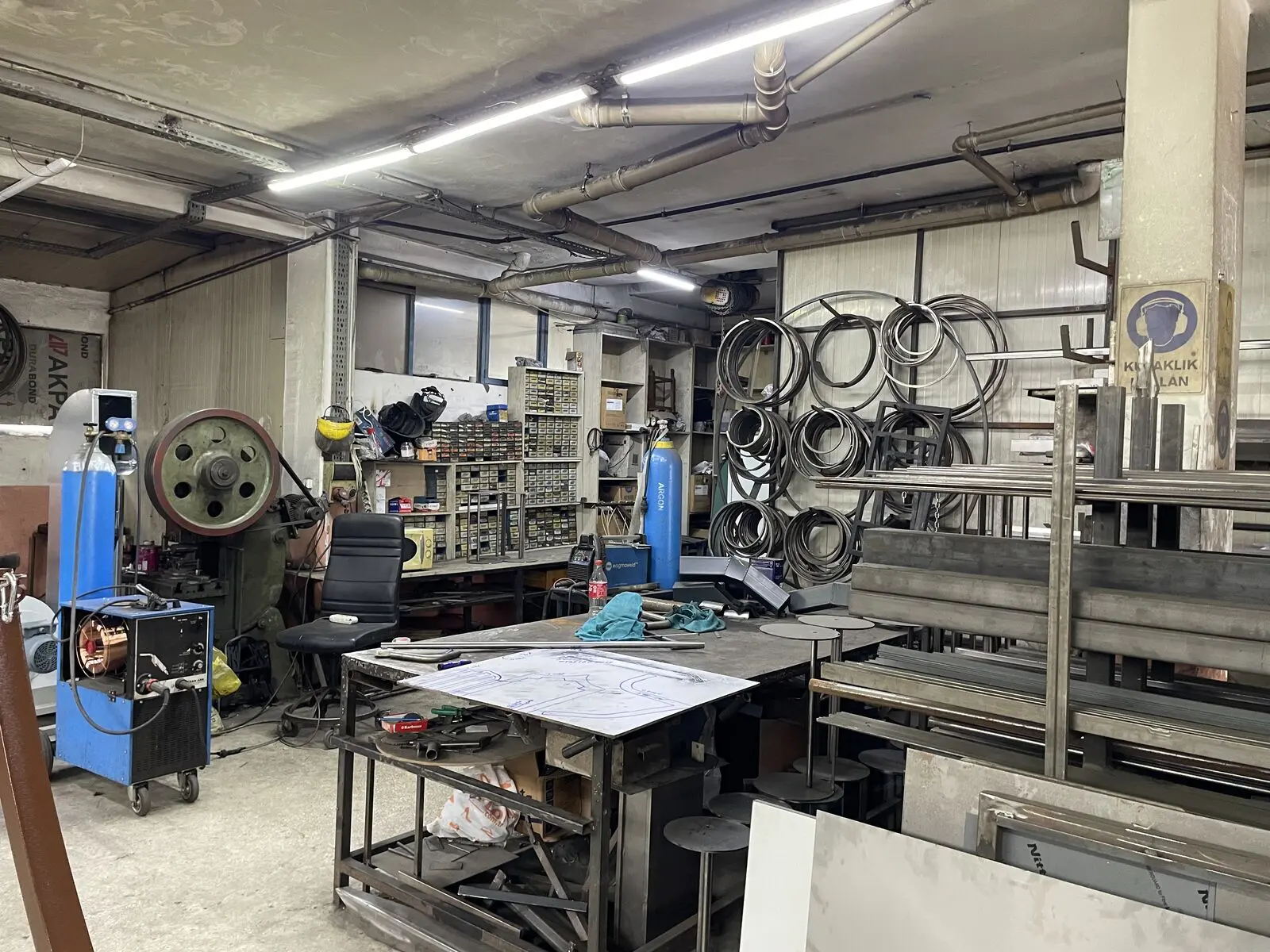 Atölyede kaynak makinesi, çalışma tezgâhı ve arka duvarda asılı bükülmüş metal halkalar